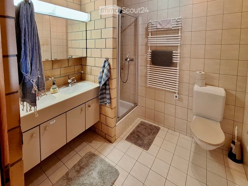 Modern bathroom with beige tiles, a double sink vanity, a shower stall, a toilet, a towel rack with towels, and a small rug on the floor.