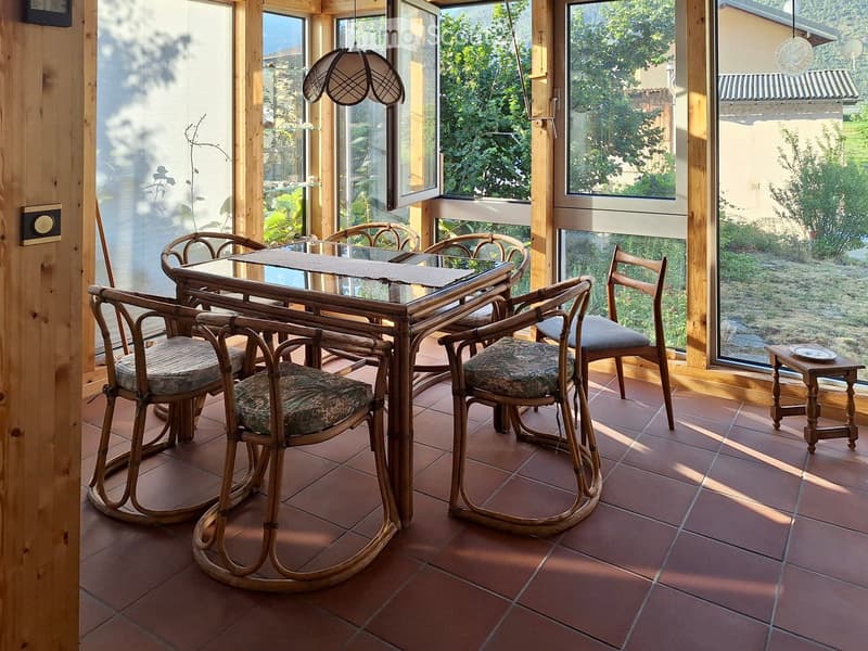 Brightly lit room with large windows, wooden furniture including a dining table and chairs, and a small side table with a plate.
