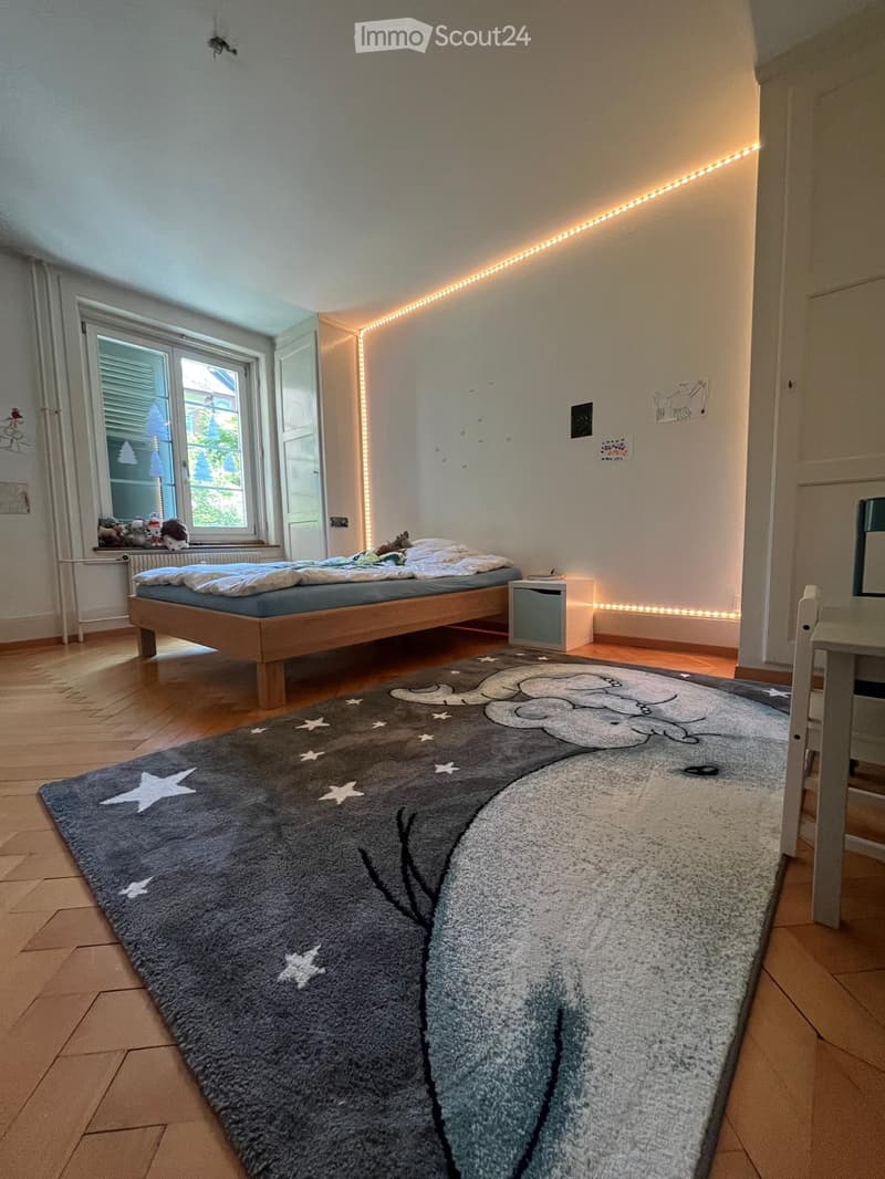 Cozy bedroom with a wooden bed, nightstand, and a large rug featuring a cloud and star design. The room has wooden flooring, white walls, and a window with blinds.