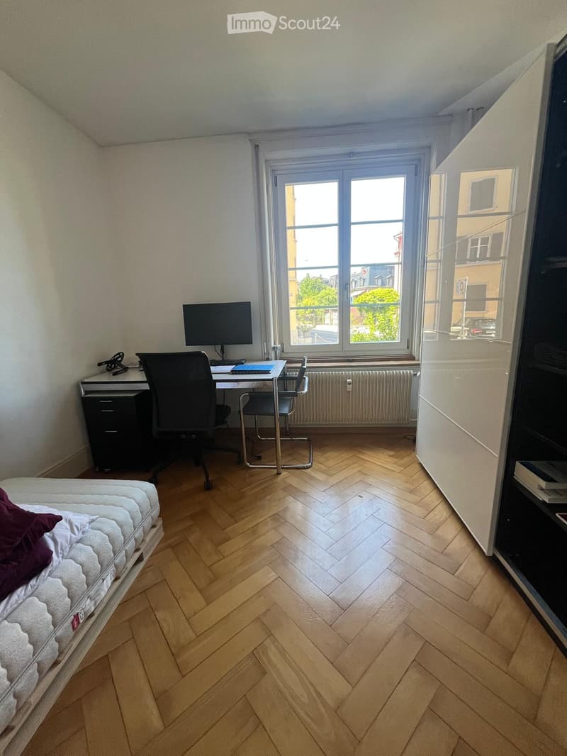 A furnished room with a bed, desk, chair, and screen, featuring wooden flooring and large windows with a view of the street.