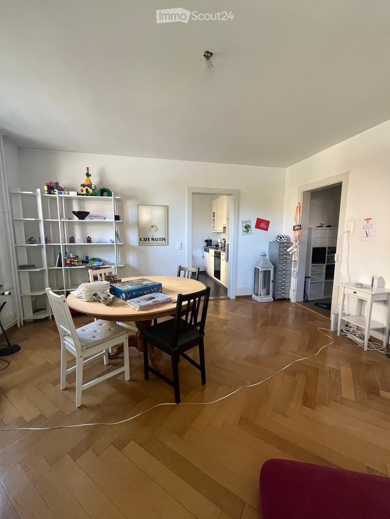 A well-lit dining area with a round wooden table, chairs, a white shelf with decorative items, and a view into a small kitchen with white cabinets, a refrigerator, and an oven.