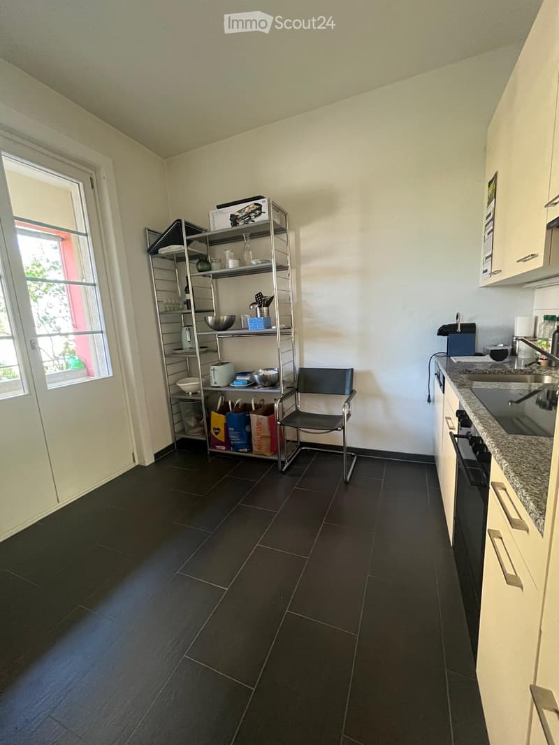 Kitchen with dark tiled floor, white walls, built-in cabinets, sink, oven, shelf with dishes, and a chair.