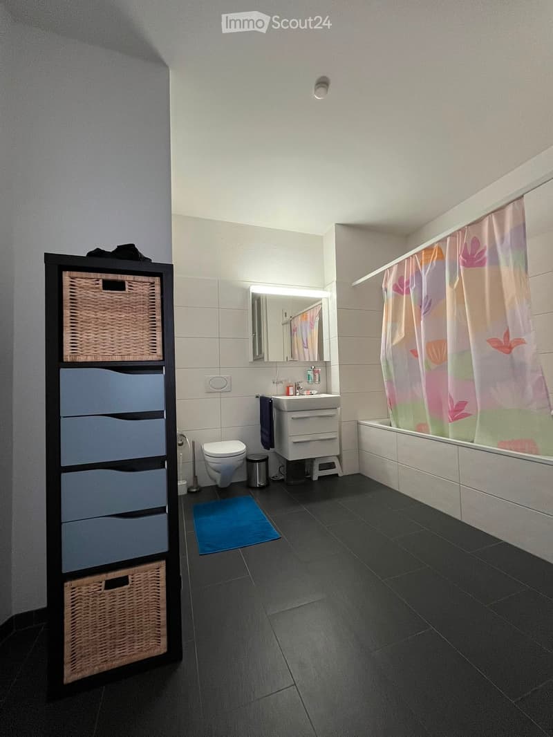 Modern bathroom with dark tiled floor, white walls, a toilet, sink with mirror, bathtub with colorful shower curtain, and a tall storage cabinet with wicker baskets and blue drawers.