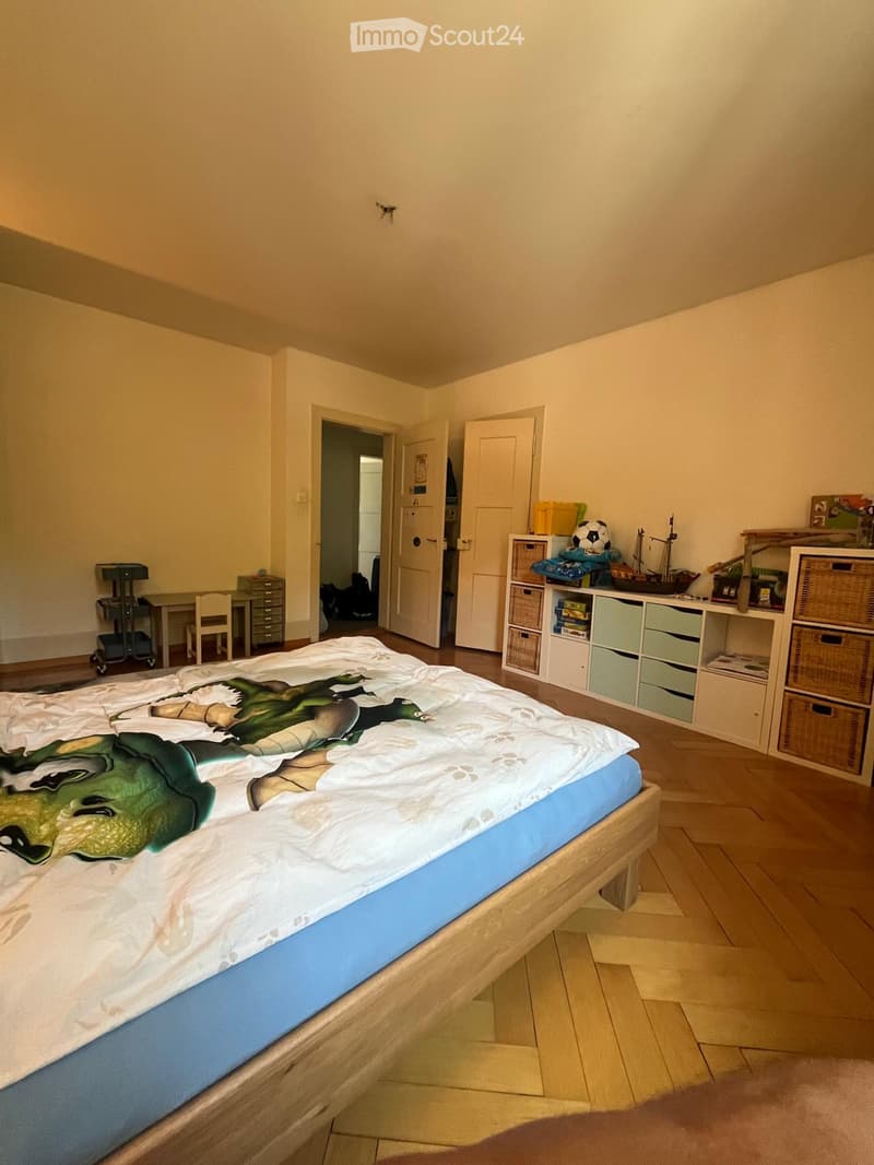 A well-lit bedroom with a large bed featuring a graphic duvet, a small table and chair, storage units, shelves with various items including a model ship and a ball, and a partially visible doorway leading to another room.