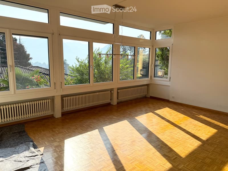 Bright, empty room with large windows offering a scenic mountain view, wooden flooring, and radiators along the walls.