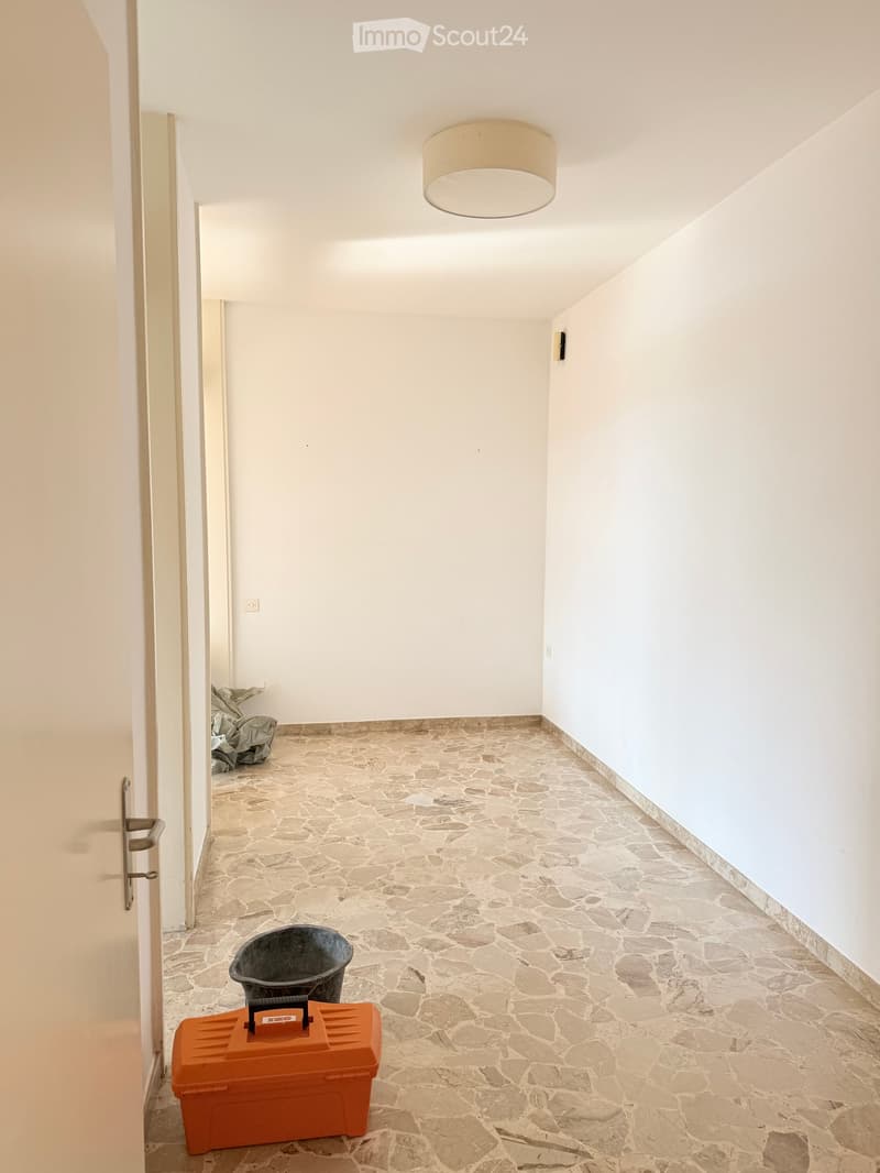 Empty hallway with stone flooring, white walls, and a single ceiling light fixture. An orange toolbox and a black bucket are placed on the floor.