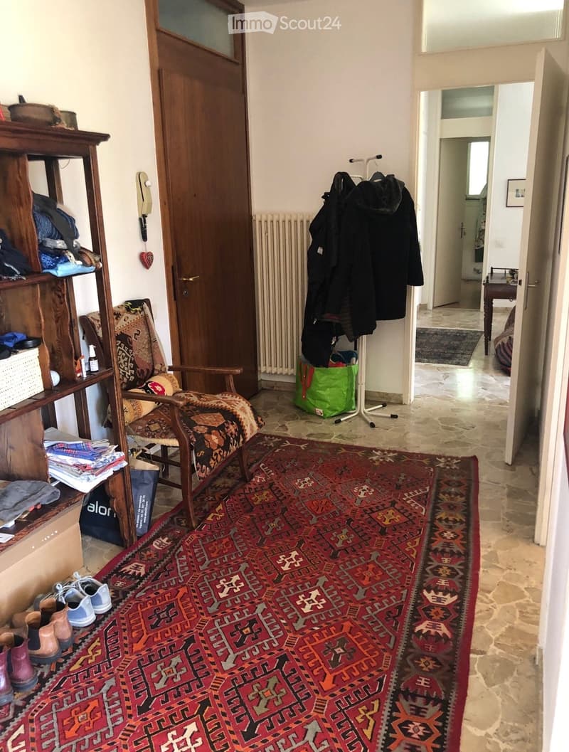 A furnished room with a patterned rug, a wooden chair with a cushion, a wooden shelf holding folded clothes and shoes, a coat rack with jackets, and a radiator. The room has stone flooring and an open doorway leading to another room.