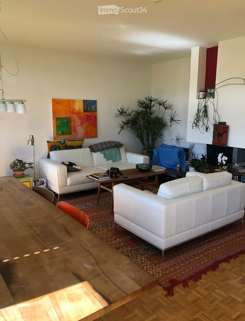Modern living room with white leather sofas, wooden coffee table, dining table, colorful artwork, and plants.