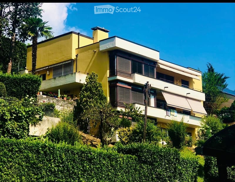 A modern two-story house with yellow and white exterior, featuring balconies, large windows, and surrounded by lush greenery and trees.