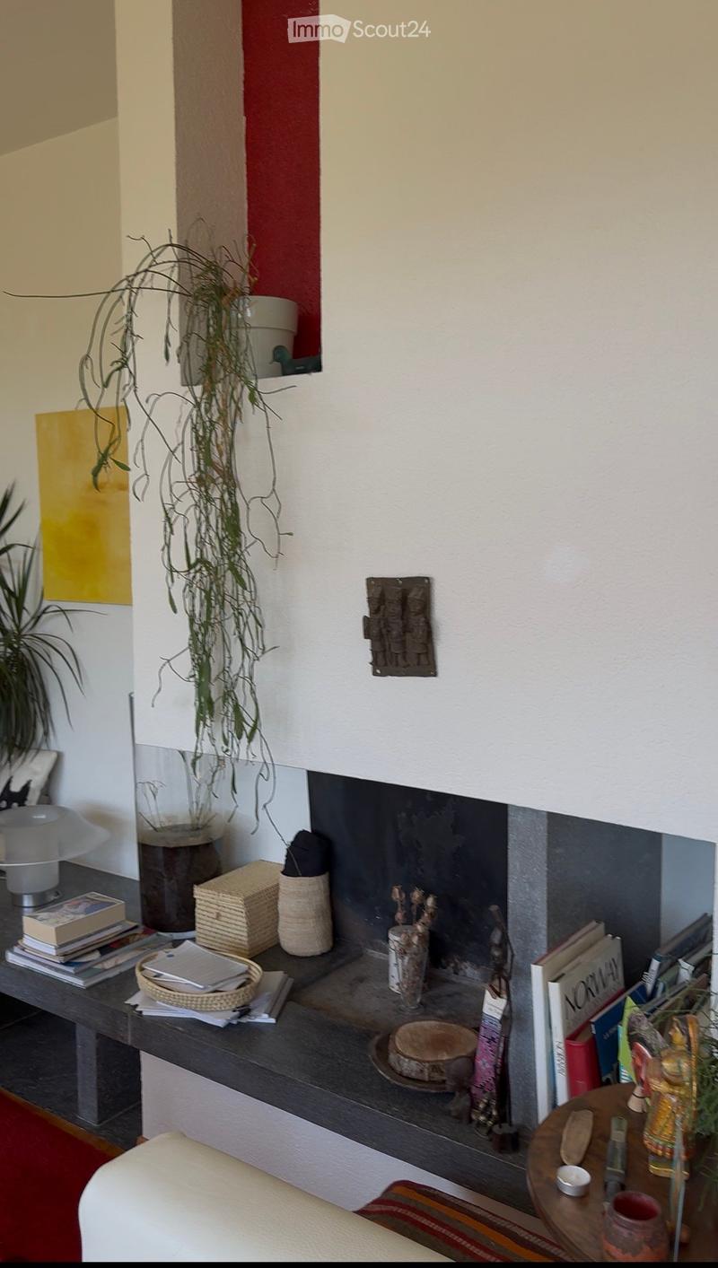 A cozy living room with a fireplace, a table with books and a lamp, a hanging plant, and decorative items on the mantel.