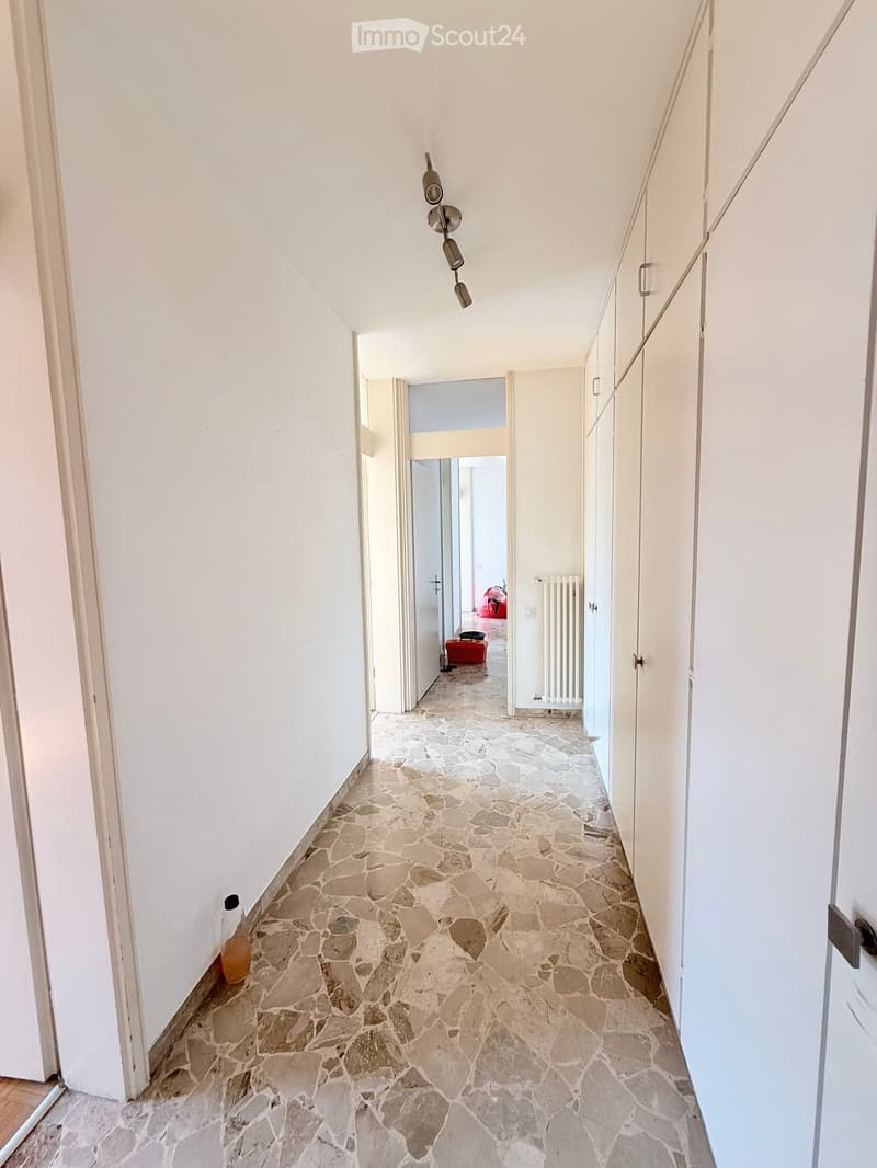 A long, narrow hallway with white walls and a stone-tiled floor. The hallway features built-in white cabinets on one side and a hanging light fixture above. At the end of the hallway, there is an open doorway leading to another room.