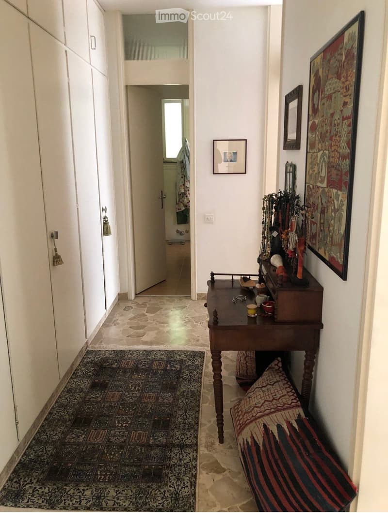 A narrow hallway with stone flooring, white walls, and a small wooden table with decorative items. The hallway features a patterned rug, framed artwork, and a doorway leading to another room.