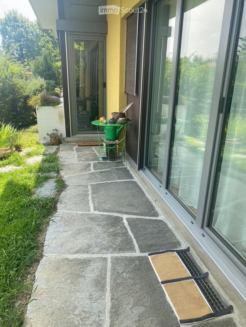 A stone-paved outdoor area adjacent to a modern house, featuring a small green metal table with decorative items, a doormat, and a small garden with potted plants.