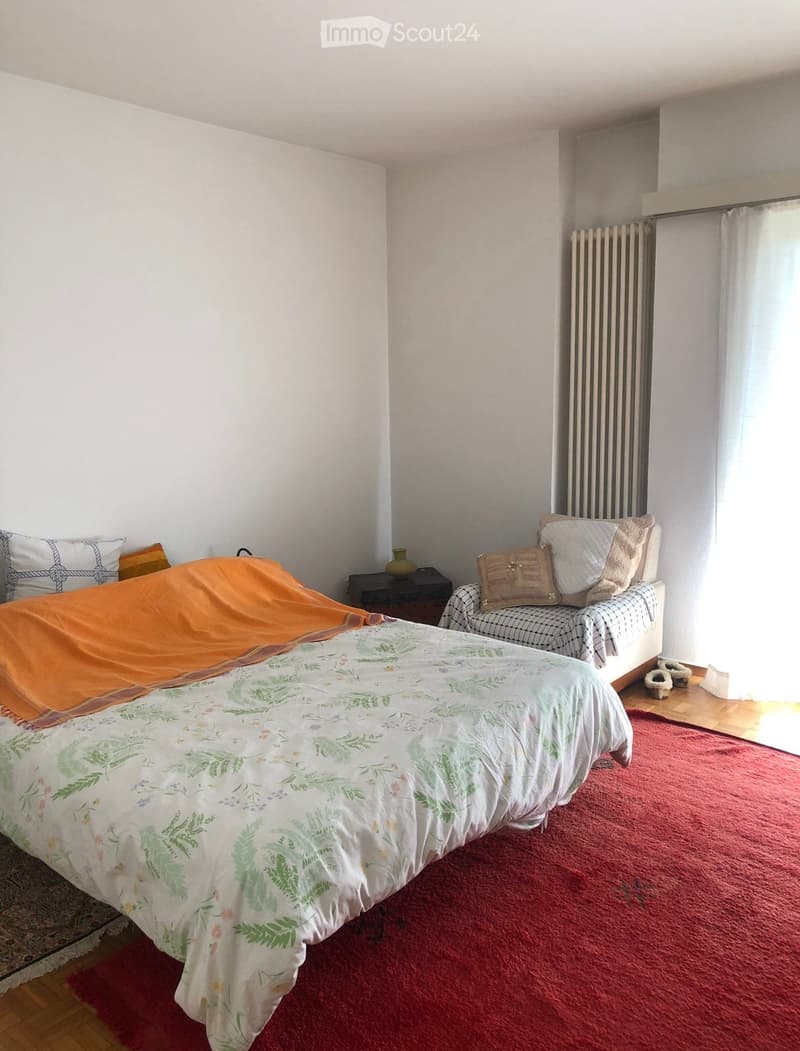 A cozy bedroom with a large bed covered in a floral duvet and an orange throw, a small sofa with patterned cushions, a nightstand with a vase, and a radiator near the window with white curtains.