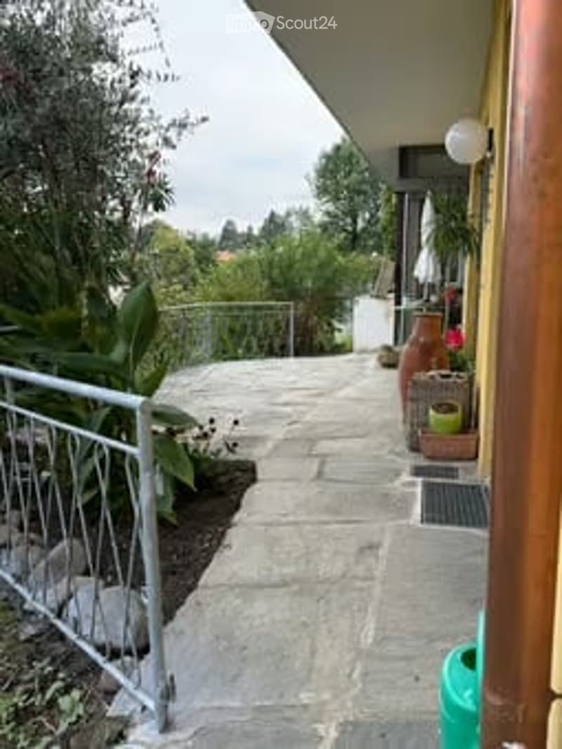 A narrow concrete pathway leads to a small balcony area, bordered by a metal railing and lush greenery, with potted plants and a small umbrella visible on the right.