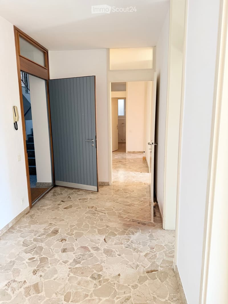 A long, narrow hallway with stone tile flooring and white walls. Features include a wooden door on the left leading to a staircase, a grey door in the center, and a partially open door on the right leading to another room.