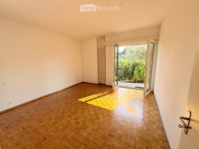 Empty room with wooden herringbone flooring, white walls, and a large sliding glass door leading to a balcony with outdoor greenery.