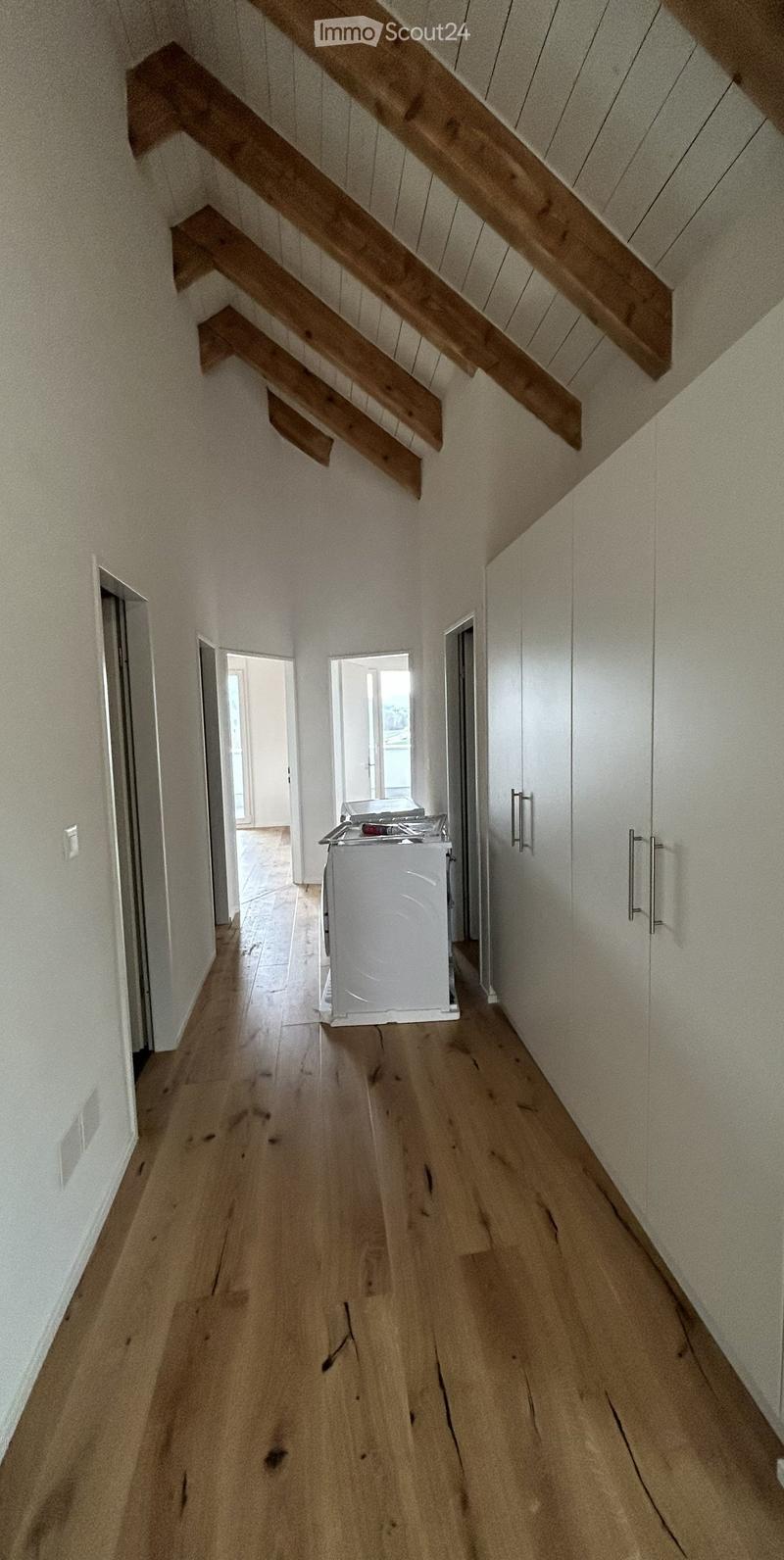 Wooden staircase with slanted ceiling and light-colored walls, leading to a hallway with wooden flooring and white walls. Laundry area visible at the end of the hallway.