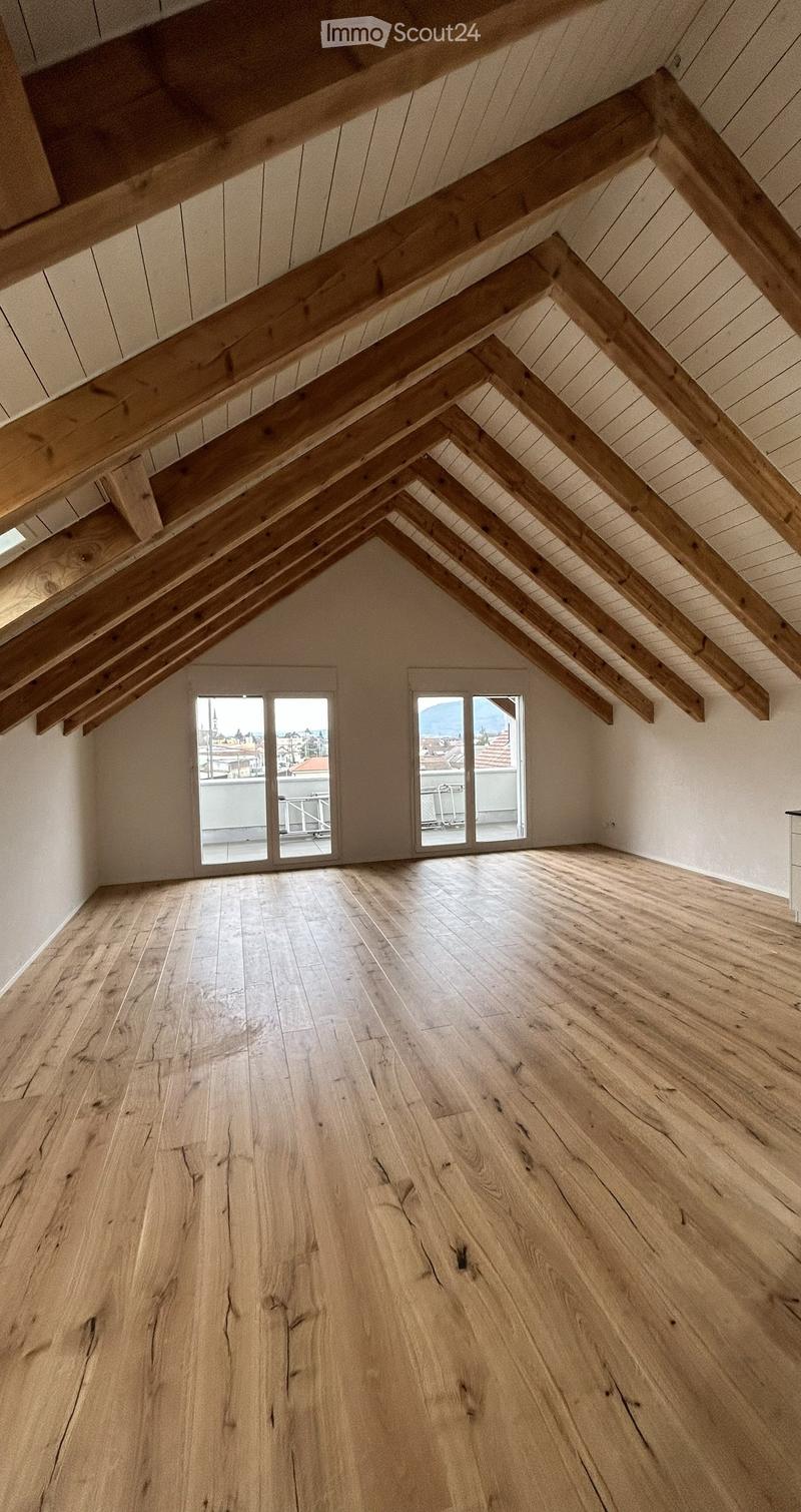 An empty attic space with exposed wooden beams, wooden flooring, and large windows offering a view of the city and mountains.
