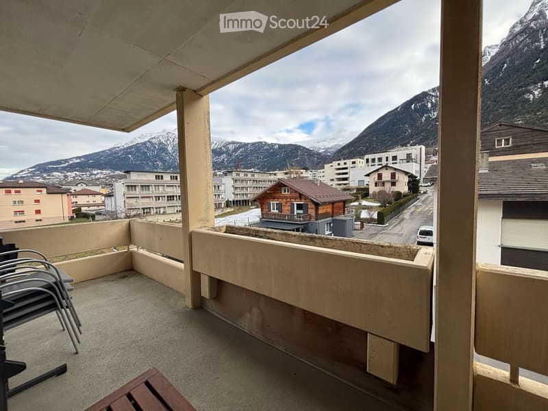 Balcony with a view of snow-capped mountains and town buildings, featuring a wooden table and metal chairs.