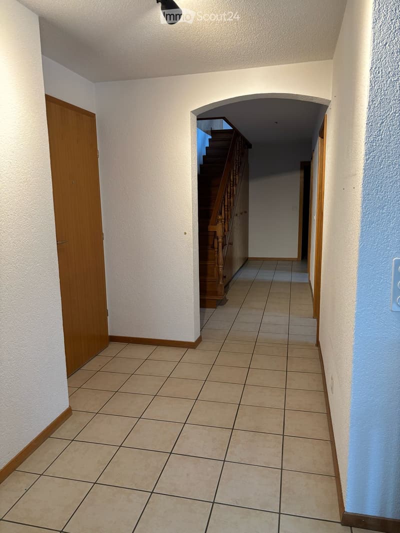 Empty hallway with tiled floor, white walls, wooden door on left, and staircase leading upwards in the background.