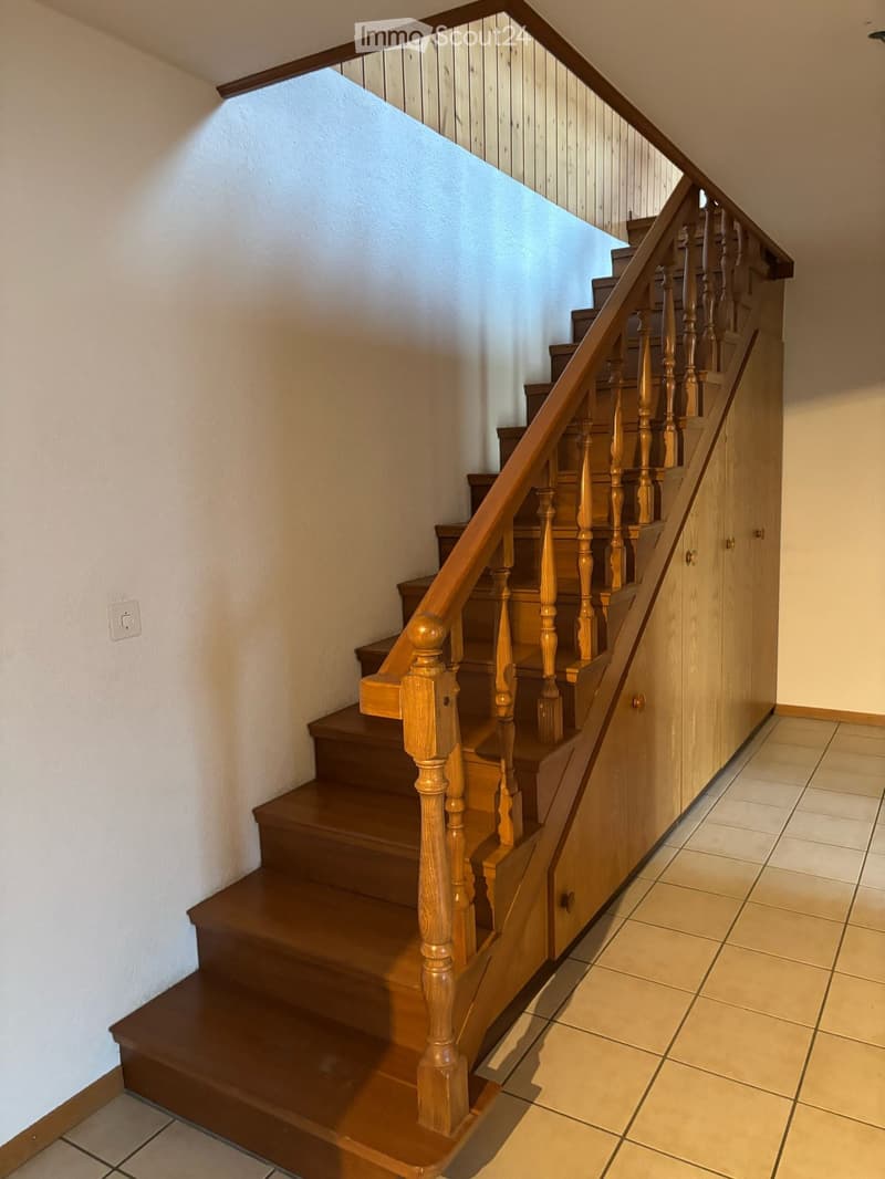 Wooden staircase with handrail and balusters, beige tiled floor, white walls, and a light switch on the left wall.