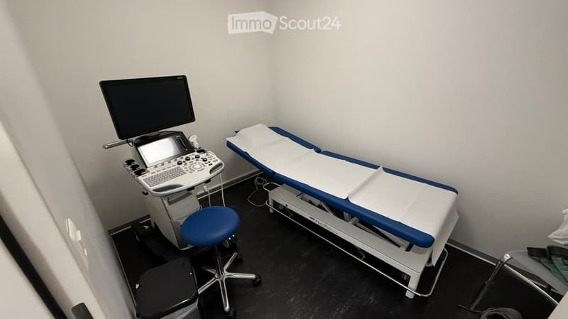 Medical examination room with an ultrasound machine, a large screen, a blue examination table, and a blue chair.