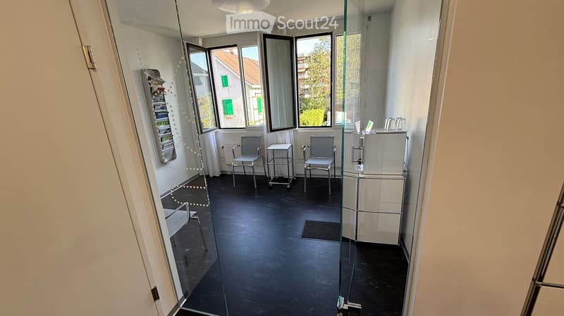 A modern, minimalistic room with large windows, black flooring, and white walls. Contains a small table, two chairs, a refrigerator, and a small cabinet.