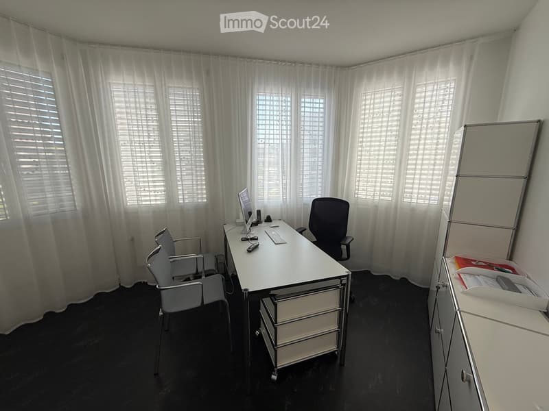 Office space with a white desk, two chairs, a computer, keyboard, mouse, and a screen. Large windows with blinds and sheer curtains.