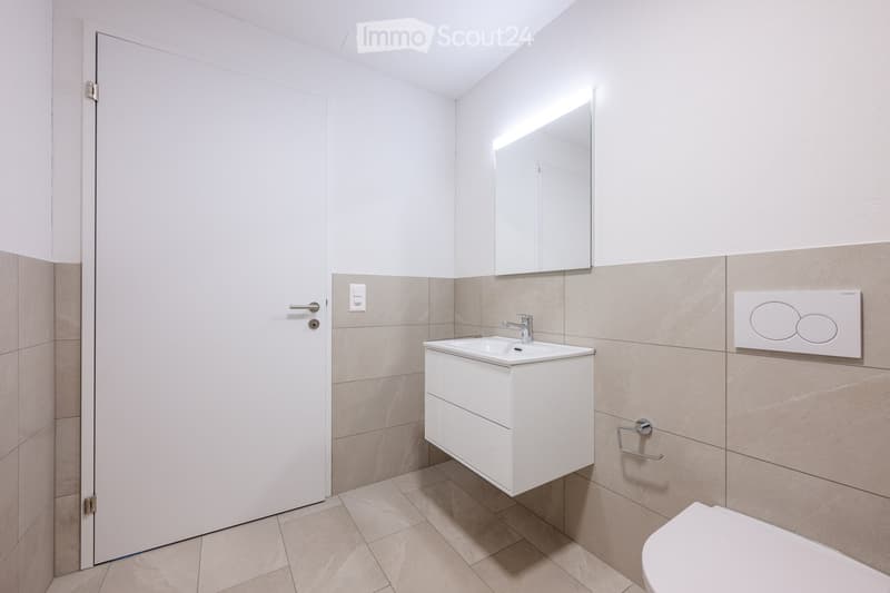 Modern bathroom with a white sink, mirror, and toilet. Beige tiles on walls and floor, white cabinet under sink, and a towel rack on the wall.