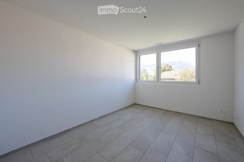 Empty room with white walls, tiled floor, and large windows offering a view of trees and mountains.