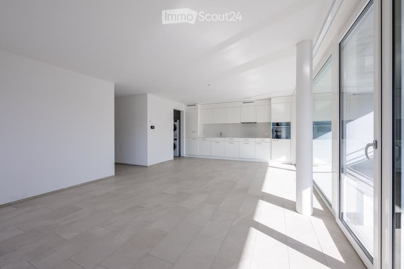 Empty room with white walls, tiled floor, large windows, kitchen with white cabinets, and a washing machine.