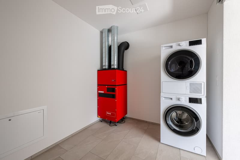 A small room with a white wall, tiled floor, a red appliance with two pipes, and a white washing machine.