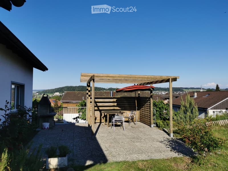 Outdoor patio area with wooden pergola, seating area, and barbecue, surrounded by greenery and distant houses.