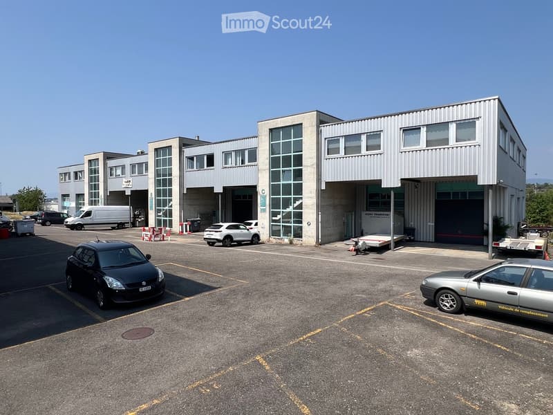 Modern industrial building with large garage doors, multiple windows, and a spacious parking lot with several cars.