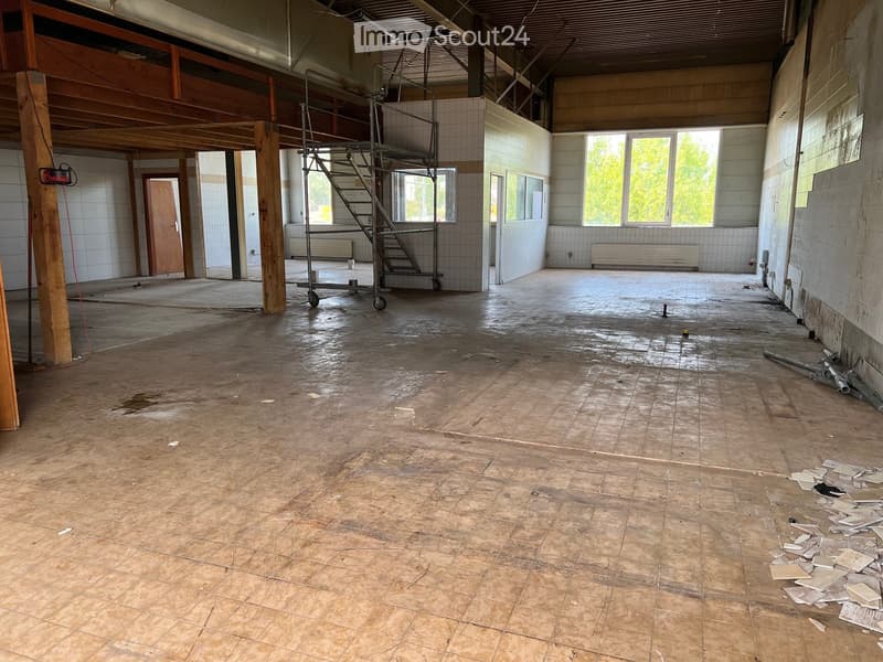 Empty room with exposed wooden beams, tiled walls, and scattered debris including broken tiles and pipes. The floor is made of concrete tiles, and there is a metal staircase leading to an upper level.
