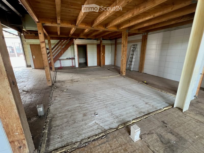 Empty room with exposed wooden beams and a wooden staircase leading to another level. The floor is unfinished with visible wooden planks and concrete patches.