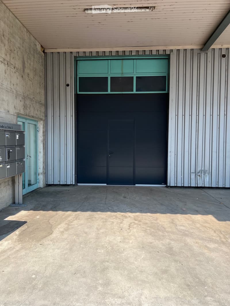 A covered garage area with a large, dark green garage door and a smaller side door, concrete floor, and a covered walkway.