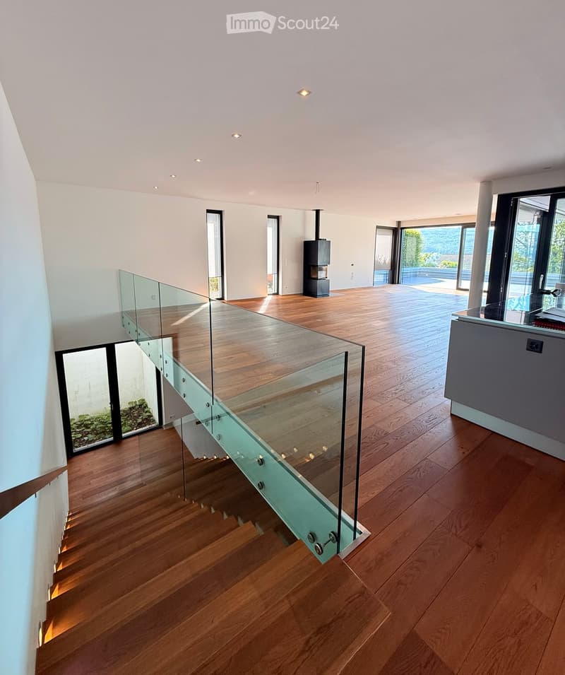 Modern hallway with wooden flooring, glass railing, and recessed lighting. Features large windows and a view of the outdoor area.