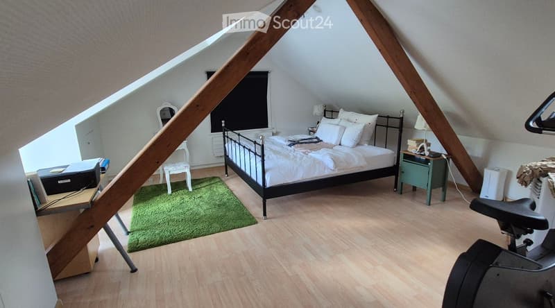 Cozy attic bedroom with wooden beams, slanted ceilings, and skylight windows. Features a metal-framed bed, nightstand with lamp, small desk with laptop, and a green rug.