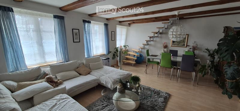 A modern living room with a large sectional sofa, glass coffee table, dining area with chairs, and decorative plants. The room features wooden beams on the ceiling and large windows with sheer curtains.