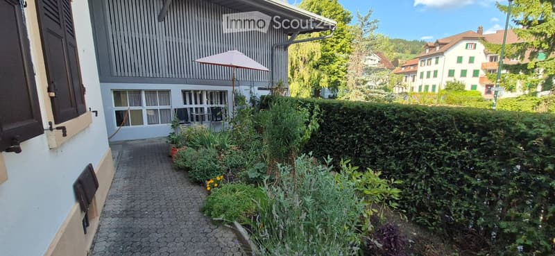 A narrow garden path lined with greenery and potted plants leads to a building entrance. The building has a modern facade with large windows and a balcony area with an umbrella. The surrounding area is lush with trees and other houses visible in the backg
