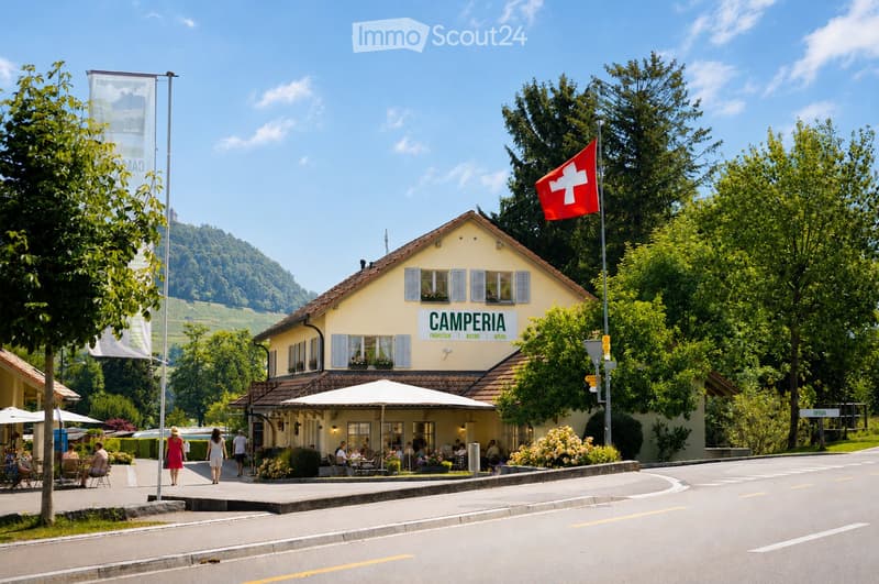 A yellow building labeled "CAMPERIA" with a Swiss flag, surrounded by trees and a scenic hillside in the background. The building has a restaurant area with outdoor seating and umbrellas.