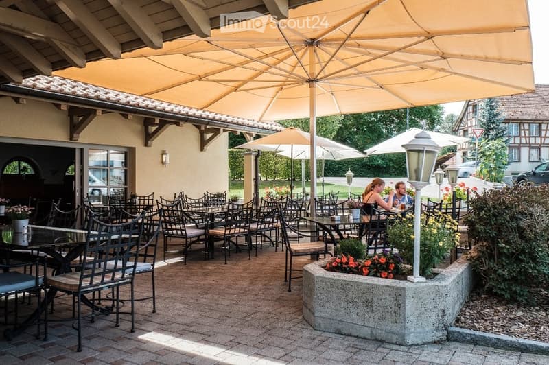 Outdoor seating area with tables and chairs under large umbrellas, surrounded by plants and flowers, with a building in the background.
