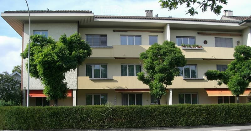 A three-story residential building with white and beige exterior, multiple windows, balconies, and surrounding greenery.