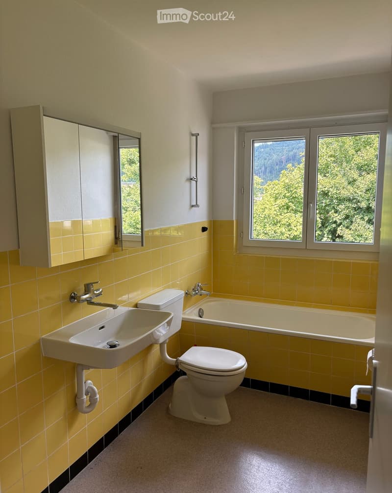 Bright yellow-tiled bathroom with a white sink, toilet, bathtub, and mirrored cabinet. Large windows offer a view of greenery outside.