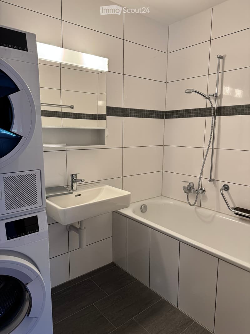 Modern bathroom with white tiles, bathtub, sink, showerhead, and a private washing machine.
