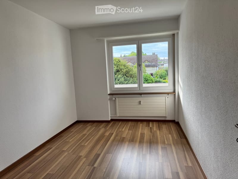 Empty room with wooden flooring, white walls, and a large window offering a view of the outside. A radiator is visible below the window.