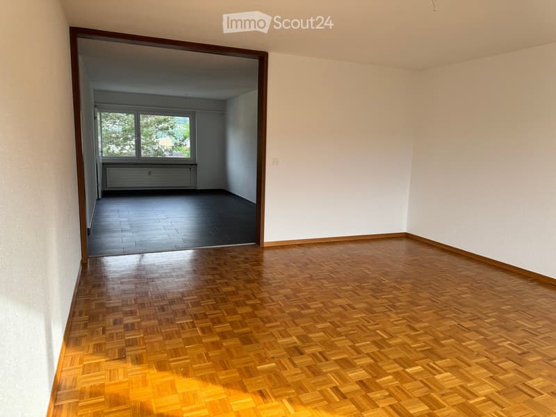 Empty room with wooden flooring and white walls, featuring an open doorway leading to another room with dark flooring and windows.