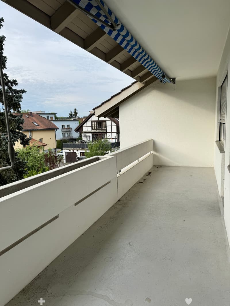 Balcony with white walls, concrete floor, and a blue and white striped awning extending from the roof.